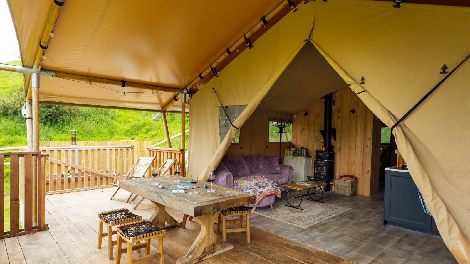 The interior of the cozy Hadspen Glamping tent features wooden furniture, including a long table and benches. There's a purple sofa nestled inside, with a view of green hills through the open front. The decor is warm and inviting, perfect for a relaxed retreat.