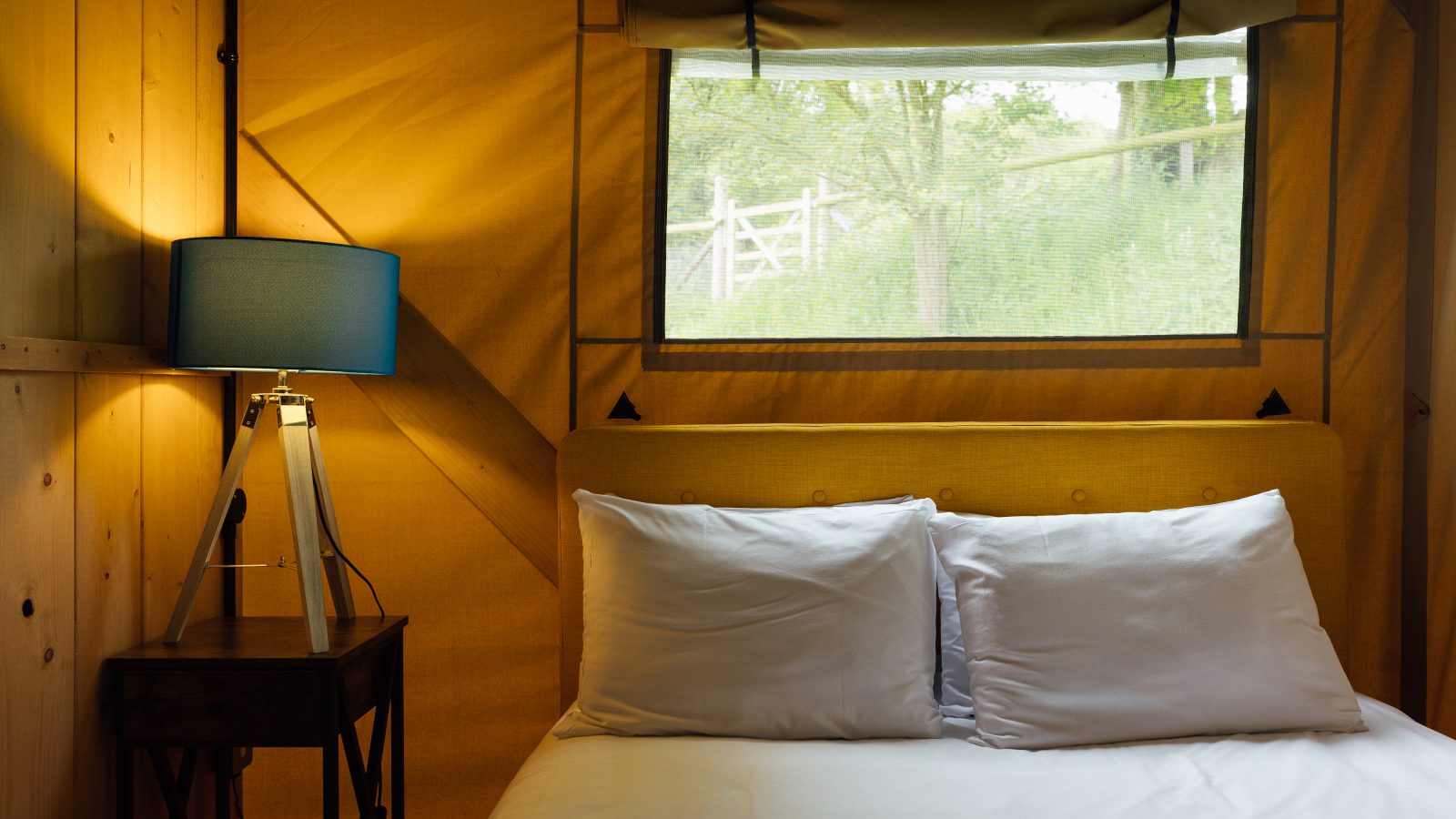 Cozy bedroom inside a glamping tent cabin in Hadspen, featuring a neatly made bed with white pillows and sheets. A small wooden nightstand holds a tripod-style lamp with a blue shade. Soft natural light filters through a window, revealing greenery outside.