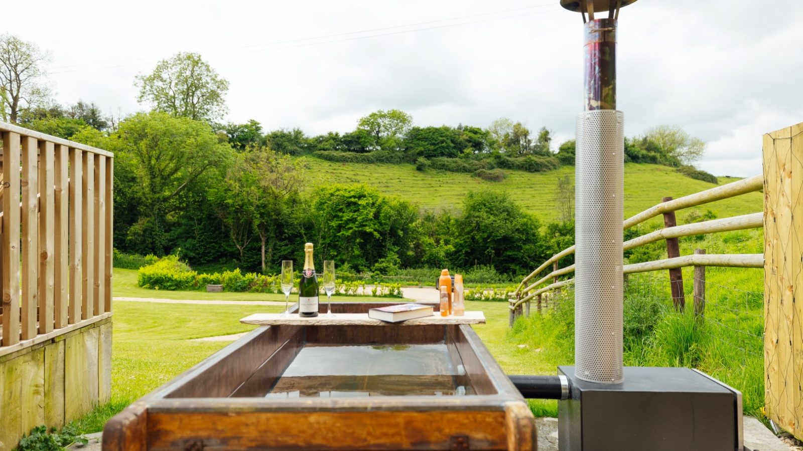 A wooden hot tub with a wood-burning stove is set outdoors at Hadspen Glamping, surrounded by lush greenery and rolling hills. A bottle of champagne, two glasses, and sunblock rest on a table beside the tub, under a partly cloudy sky.