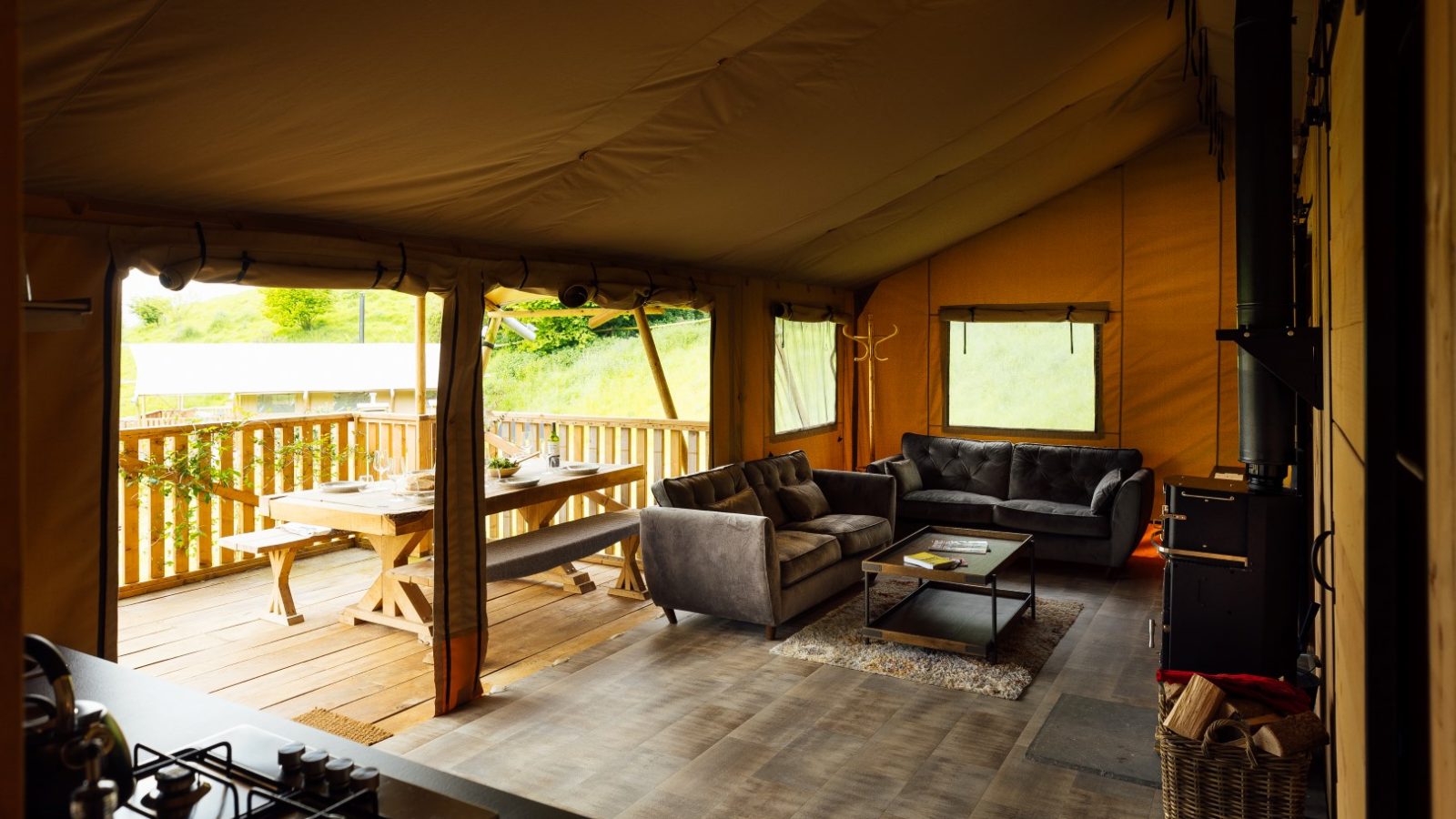 Nestled in the serene surroundings of Hadspen, this cozy glamping tent features a living area with gray sofas, a wooden coffee table, and a stove. Outside on the deck, a dining table awaits under a sunny sky, enveloped by lush greenery.