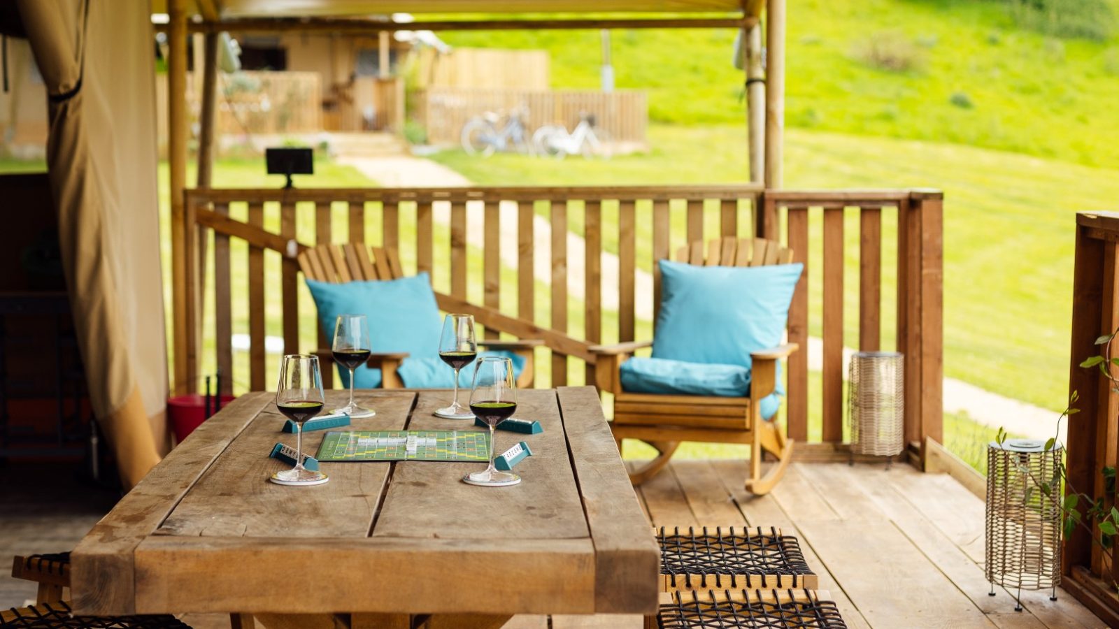 A cozy and inviting wooden deck perfect for a glamping escape. The table is set for a game with a board, wine glasses, and cards. Two chairs with blue cushions face the table, overlooking the green lawn and the blurred backdrop of Hadspen Glamping's enchanting charm.