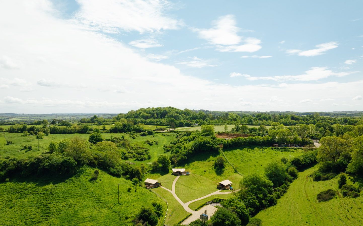 Aerial view of Hadspen Glamping, nestled in a lush green landscape with rolling hills and meadows under a partly cloudy sky. A few scattered buildings dot the scenery, with a winding road elegantly cutting through the greenery.