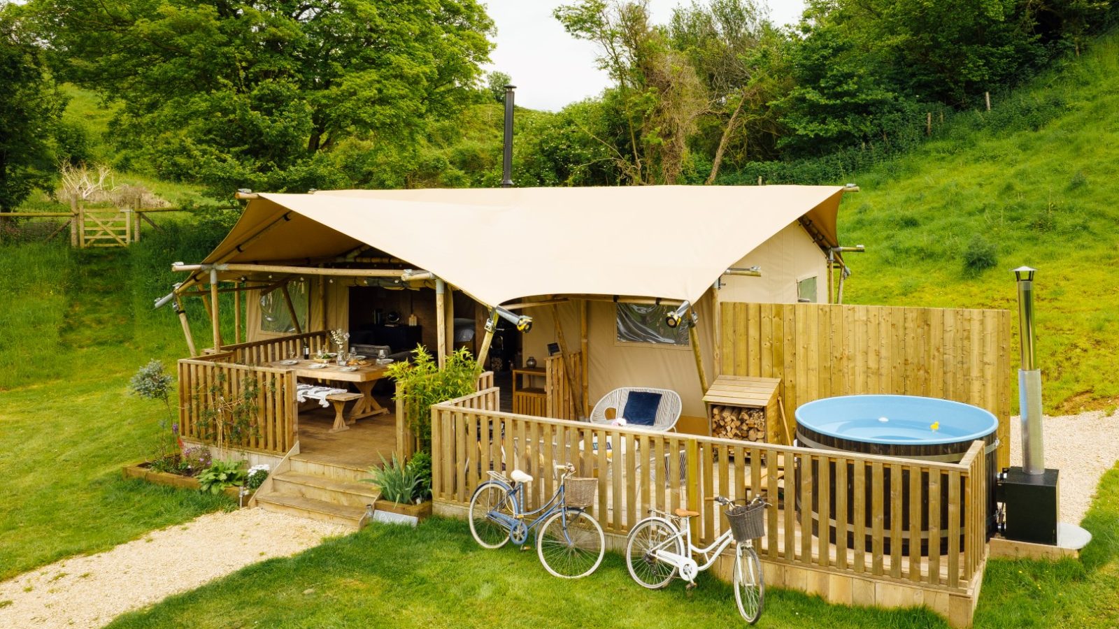 Set in the lush green landscape of Hadspen, this luxury glamping tent features a canvas roof, a wooden deck with a dining area, and a fenced area hosting a round hot tub. Two bicycles are parked nearby, offering an invitation to explore the surrounding trees and grassy hills.