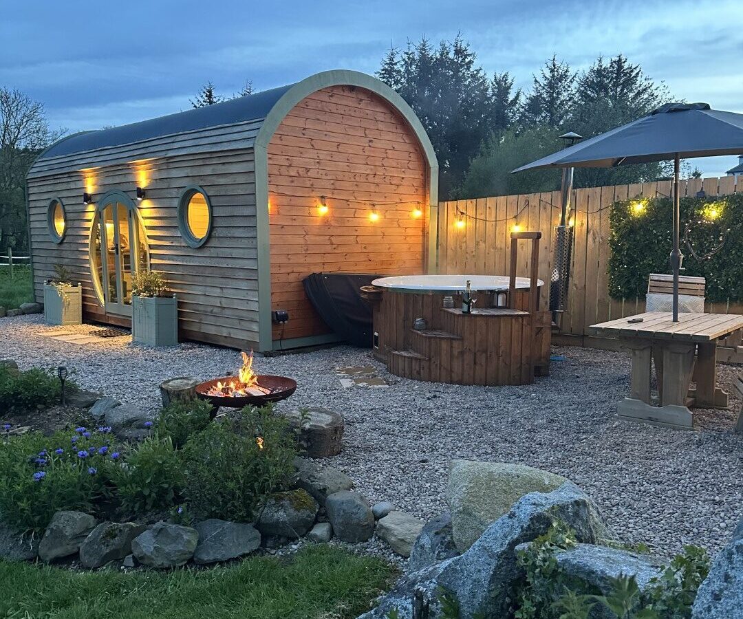 Experience the charm of Freedom Fields in this cozy outdoor scene, featuring a wooden pod perfect for glamping. Relax with a hot tub, fire pit, picnic table, and umbrella on a gravel area surrounded by lush greenery.