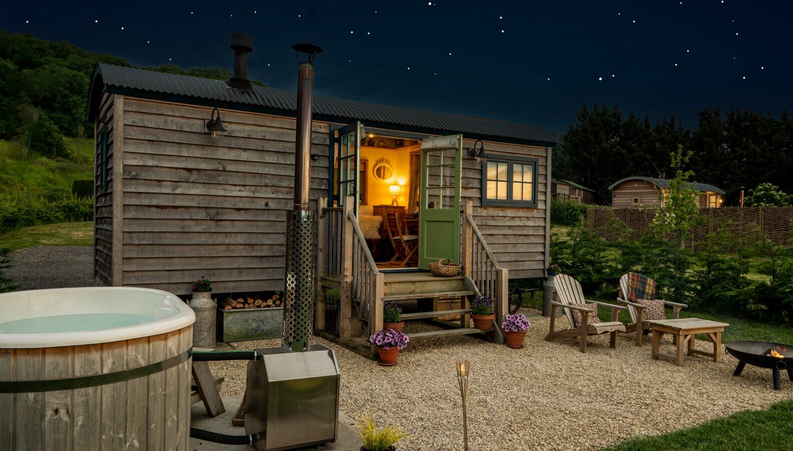 A cozy wooden cabin resembling charming huts, with a lit interior, surrounded by chairs, a hot tub, and a gravel yard, under a starry night sky.