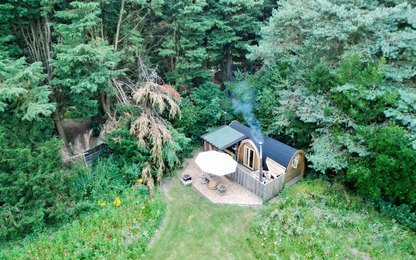 Aerial view of a small cabin with a curved roof nestled among the tall trees and lush greenery of Lahtle Wood. This glamping retreat features a patio with a table and umbrella next to the cabin, surrounded by grass and wildflowers.