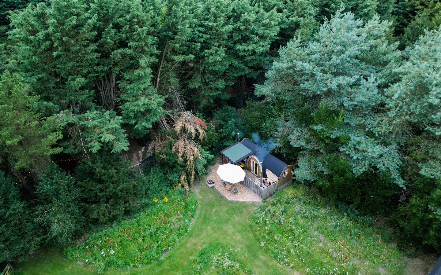 Aerial view of a charming wooden cabin with a patio, nestled in a lush Lahtle forest. The cabin boasts a green roof, and there's a white patio umbrella beside it. Dense trees surround the cabin, complemented by a grassy area adorned with colorful wildflowers, perfect for glamping enthusiasts.