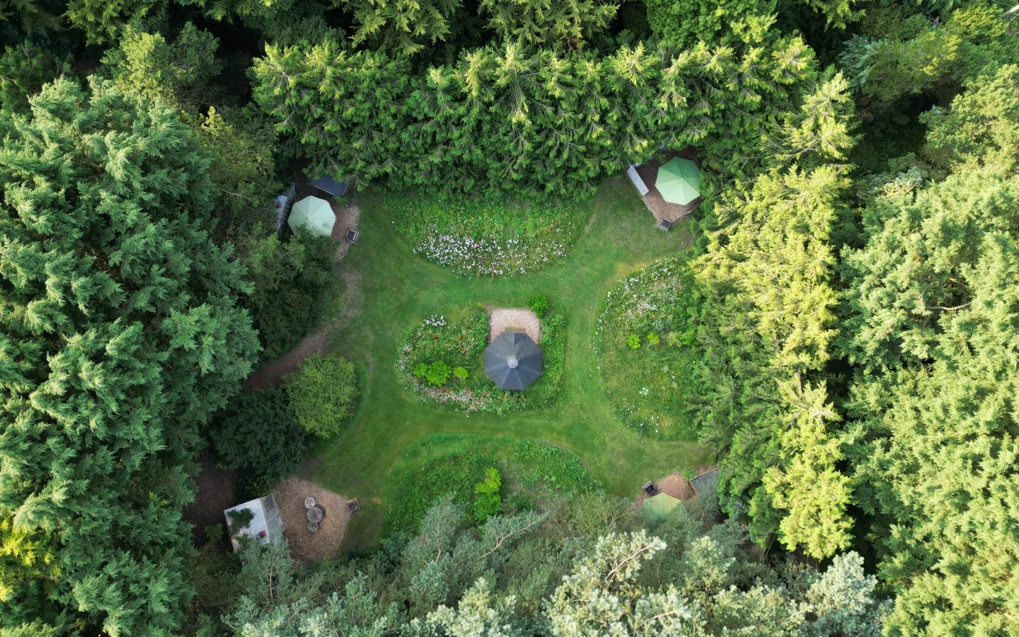 Aerial view of a lush, green forest clearing with four tents arranged in a square layout for a perfect glamping experience. The center features a garden with pathways and an umbrella, surrounded by dense evergreen trees and wood structures that blend harmoniously into the serene Lahtle landscape.