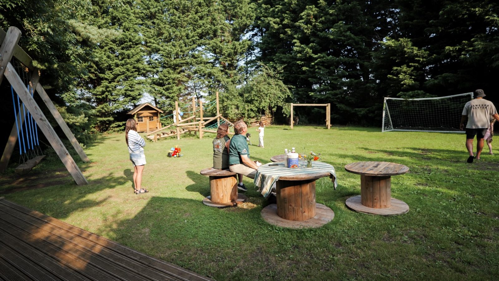 A grassy backyard at Lahtle Wood features a wooden playset and a soccer goal. Three people sit at a table with colorful cups, while others stand or walk nearby. Trees border the area, casting gentle shadows like a serene glamping retreat under the sunlit sky.