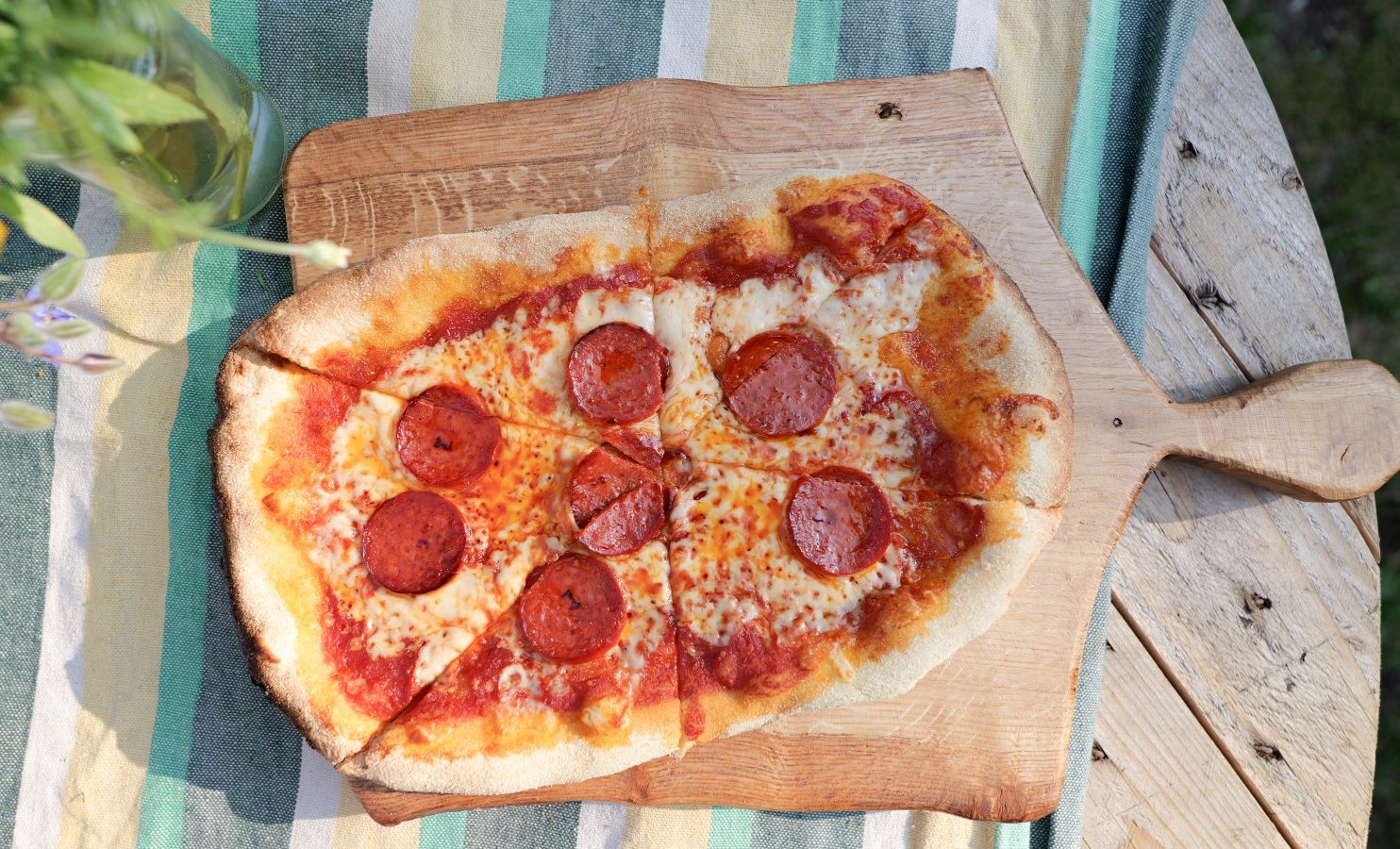 A freshly baked pepperoni pizza cut into slices, served on a Lahtle Wood board. The pizza rests on a striped tablecloth with a vase of flowers nearby. Sunlight casts shadows, enhancing the warm, rustic presentation reminiscent of glamping under the open sky.