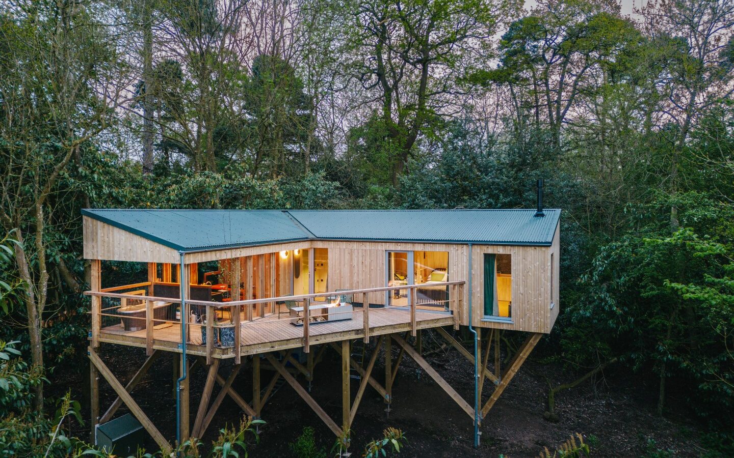 A wooden treehouse with large windows and a deck stands elevated on stilts, nestled among dense trees in a forest setting. This serene retreat invites you to rewild things and reconnect with nature in the most enchanting way.