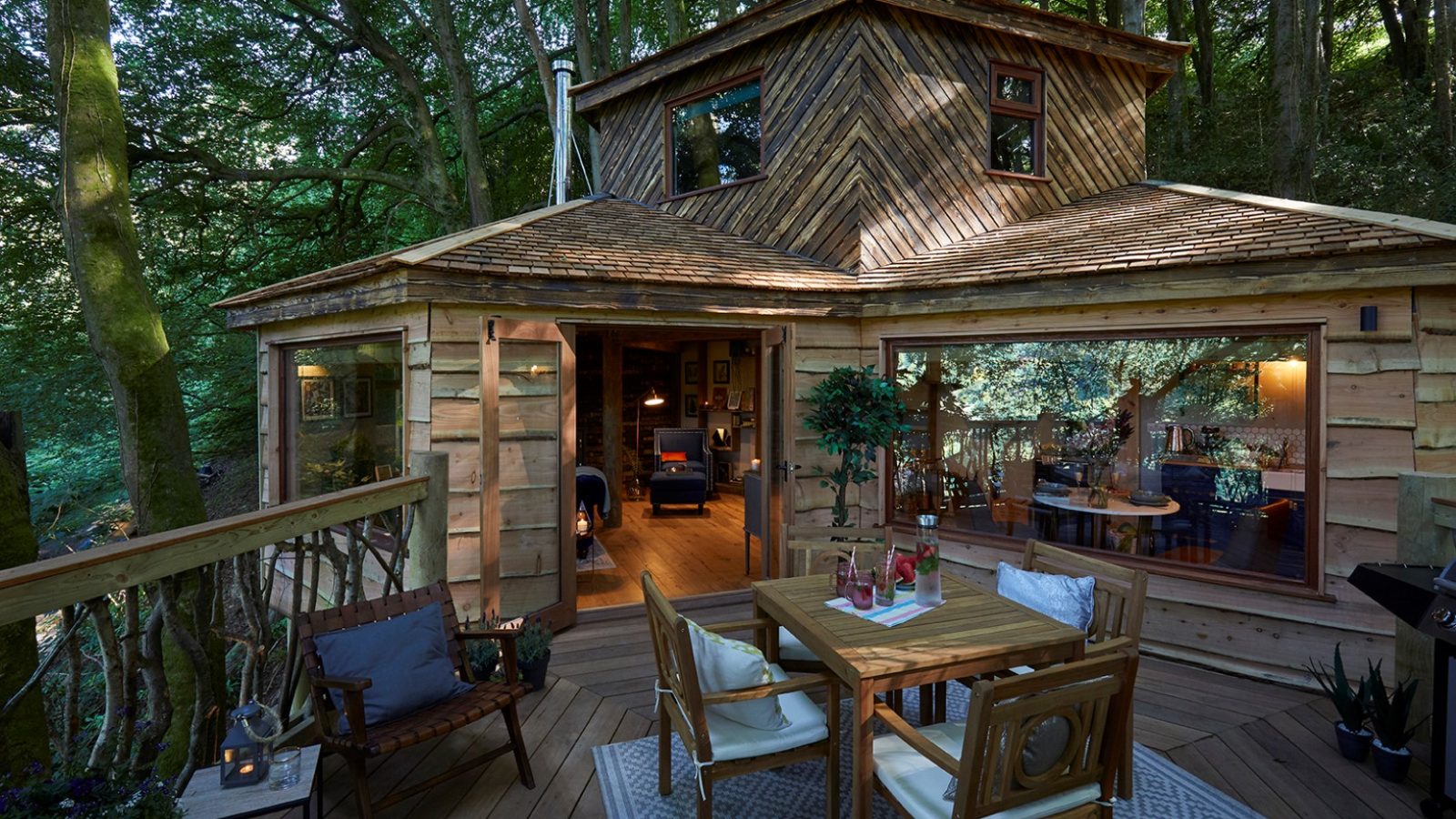 Nestled in a serene forest, this cozy wooden Ravendere Treehouse boasts large windows and offers a delightful blend of nature and comfort. The warmly lit interior reveals inviting seating with picturesque tree views, while the deck invites dining under the canopy—a perfect Ravendere accommodation.