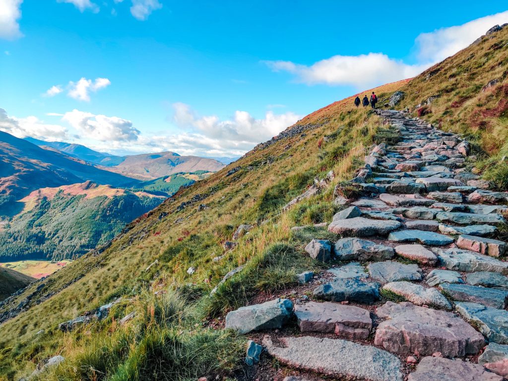 A scenic mountain path made of stone steps ascends a grassy slope, with distant hills and valleys in the background. The sky is clear with scattered clouds. Two people are walking up the path, adding a sense of scale to the expansive landscape. This route is featured in "Best British Walks - Our Top 7 Sensational Trails.