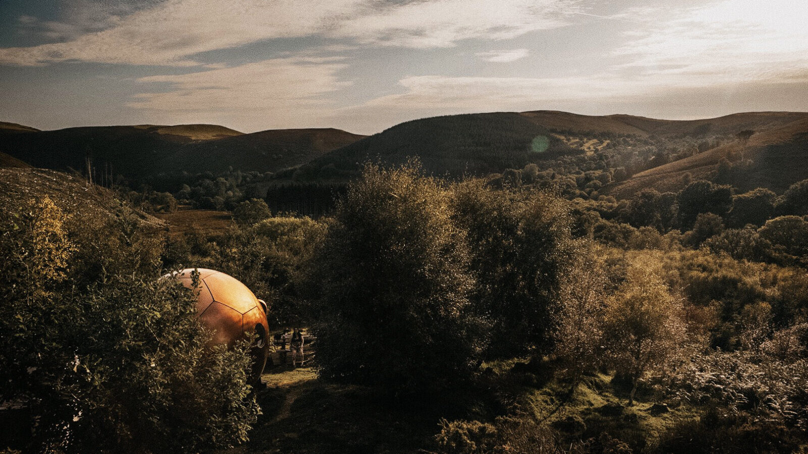 A golden dome structure is nestled among trees in the hilly Chillderness landscape, under a partly cloudy sky, offering a unique glamping experience where nature meets comfort and you might even spot a majestic Red Kite drifting overhead.