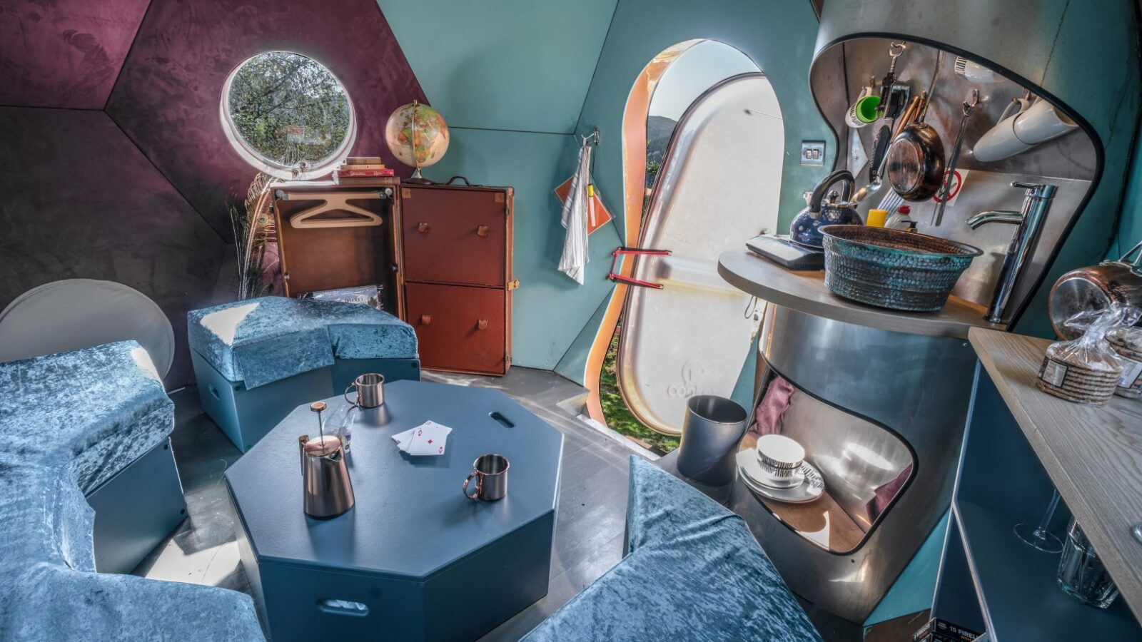 Futuristic interior of a small pod-like room designed for glamping, featuring hexagonal furniture, a circular window offering serene views, and a compact kitchenette area perfect for experiencing chillderness.