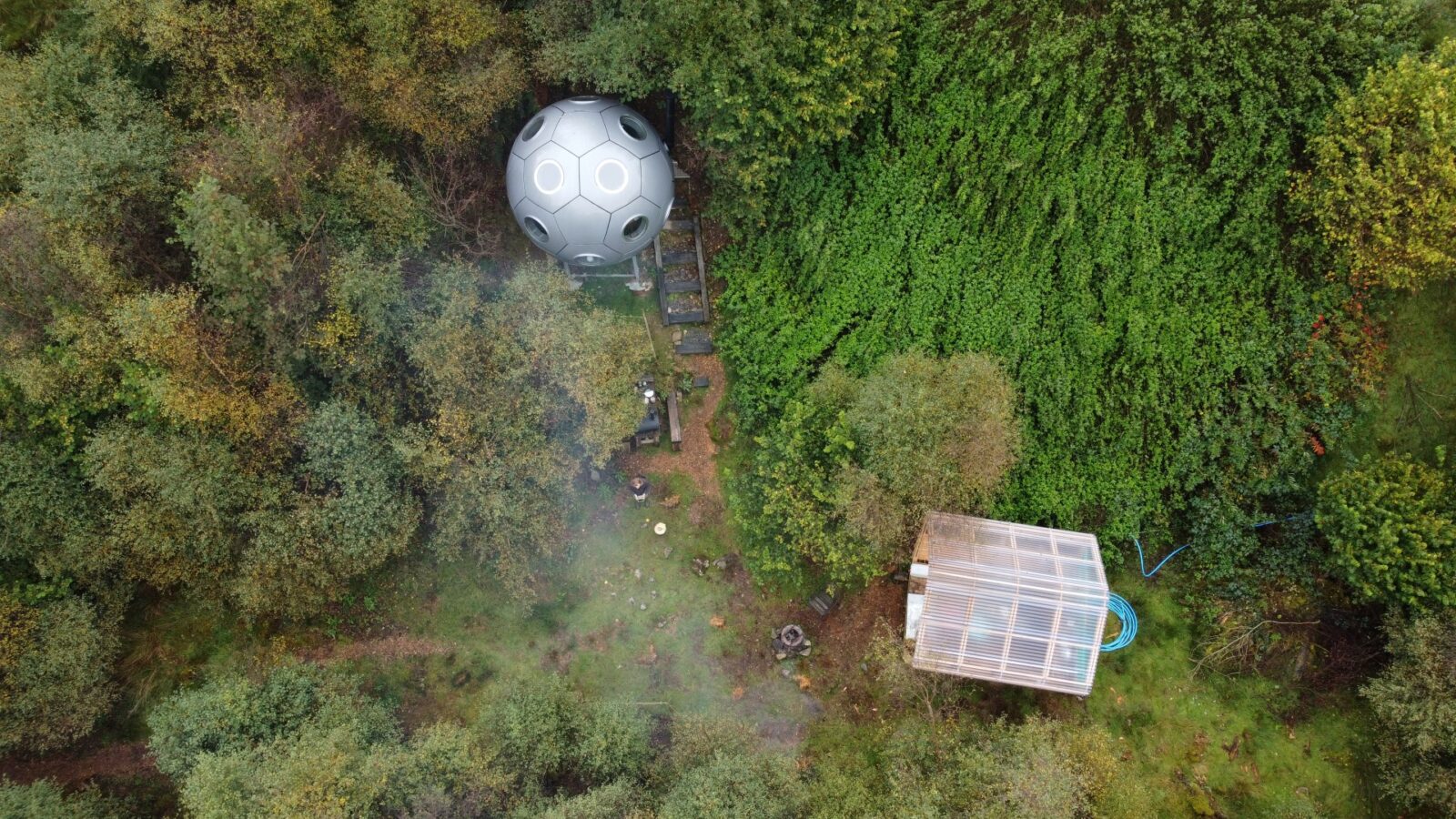 An aerial view reveals a dome-shaped structure nestled in the heart of a forest, with a small greenhouse and a grassy clearing perfect for chillderness. The serene scene is occasionally graced by the sight of a red kite soaring above.