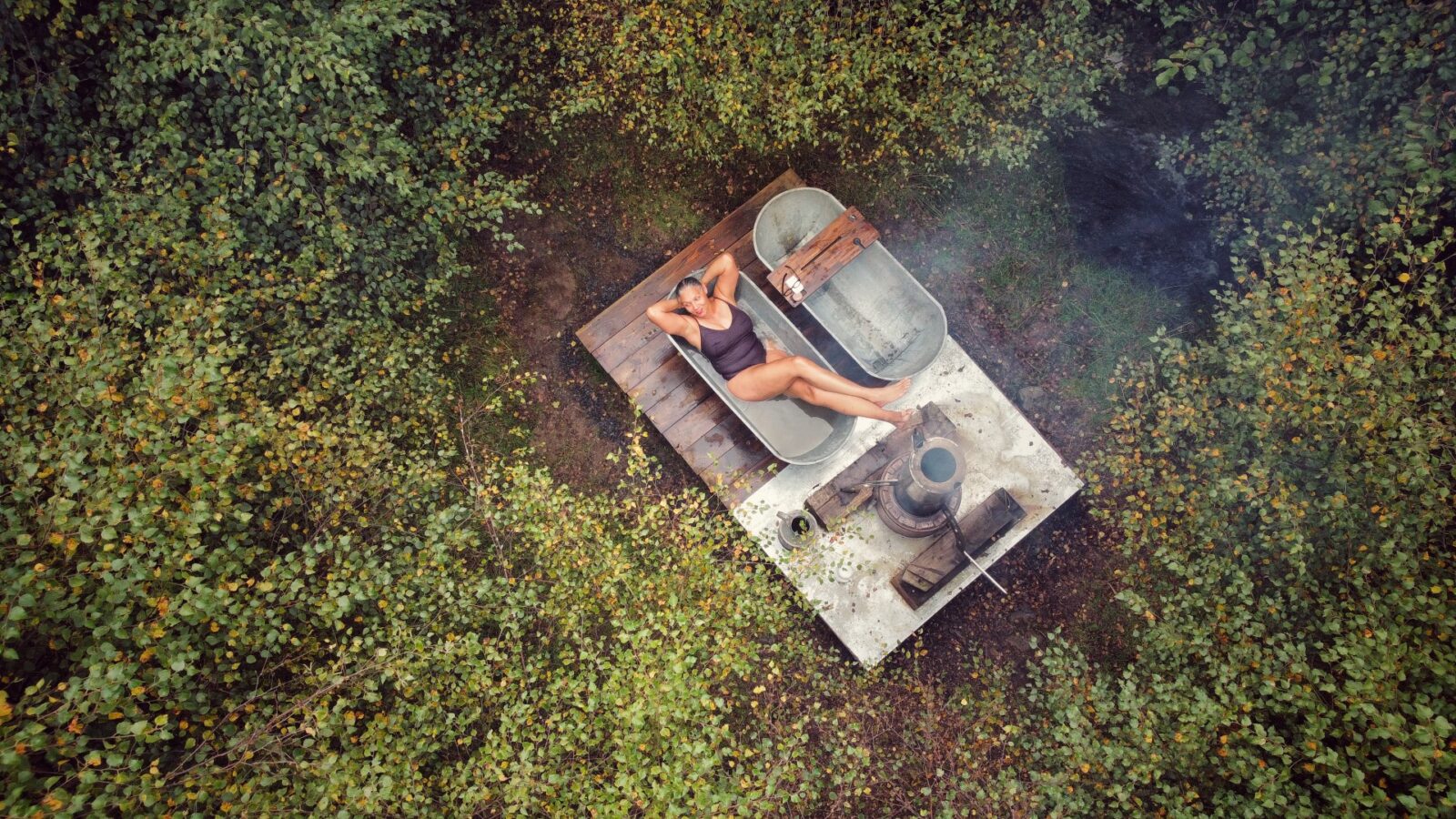 A person unwinds in an outdoor bathtub surrounded by dense, green foliage, with a rustic stove nearby, embracing the essence of chillderness at Red Kite Glamping.