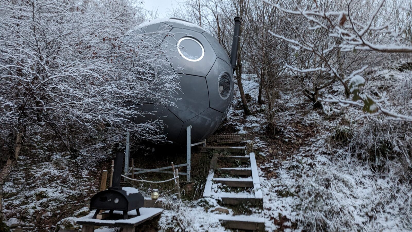 A spherical, metallic cabin with circular windows nestles on a snowy hillside, offering a unique glamping experience amidst the chillderness. Surrounded by bare trees and accessed by wooden steps, it's the perfect spot to watch a Red Kite soar overhead.