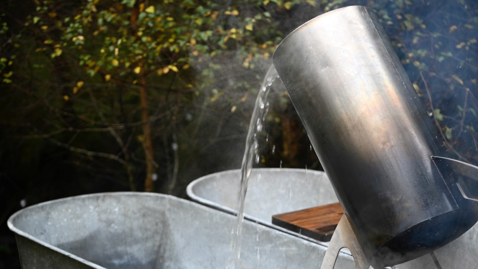 A metal container pours steaming hot water into large tubs outdoors, embracing the spirit of Chillderness. Surrounded by trees, this glamping scene invites relaxation as a Red Kite soars gracefully overhead.