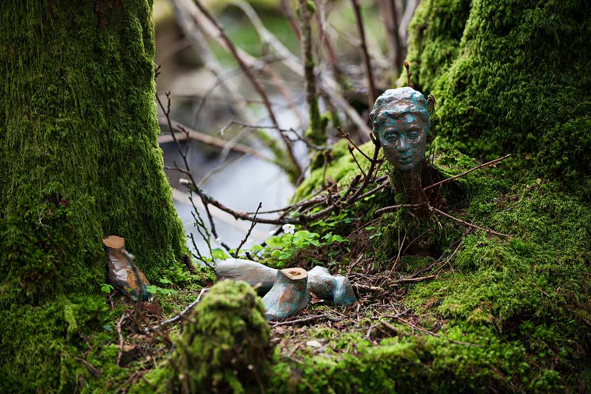 A bronze head sculpture surrounded by broken pieces rests on the mossy ground between tree trunks, offering a serene touch to the Chillderness experience.