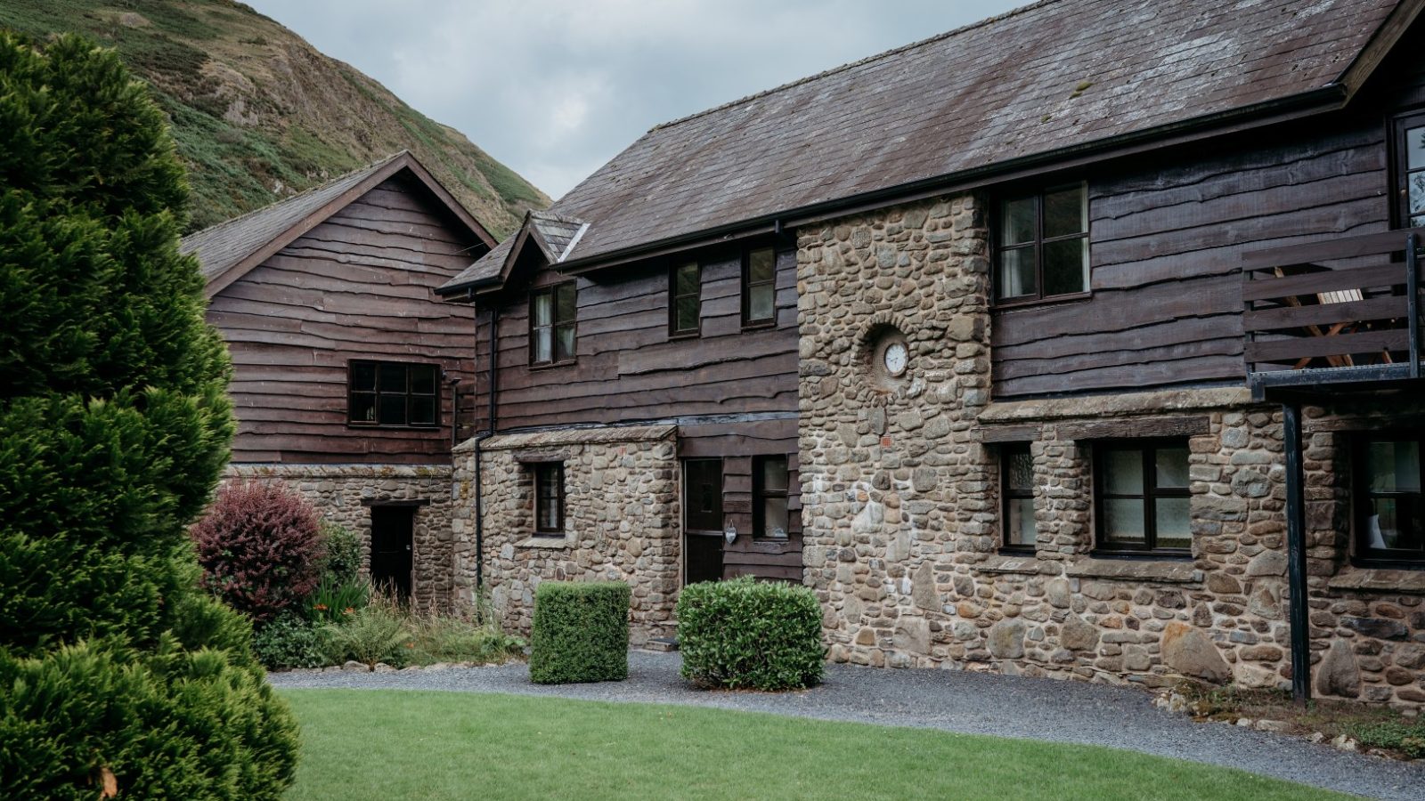 A rustic stone and wood building with two stories, reminiscent of charming Cwm Chwefru Cottages, is surrounded by green grass and shrubs. The background features a hillside under a cloudy sky, perfect for vacation rentals. Multiple windows highlight the facade, while a gravel path leads to the entrance.