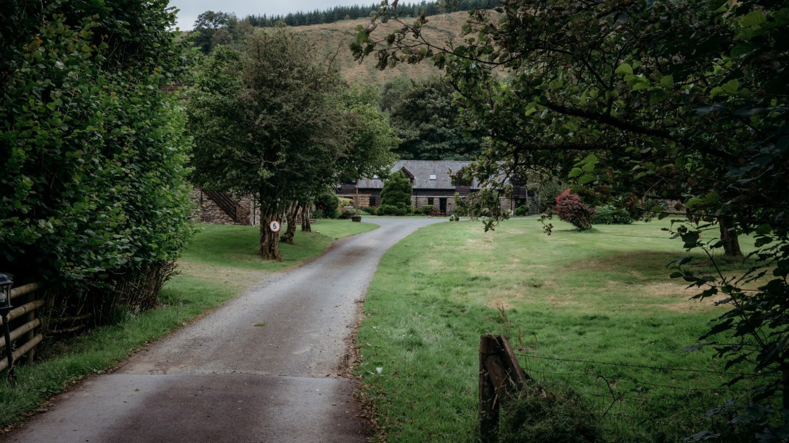 A narrow country lane leads to Cwm Chwefru Cottages, a quaint stone retreat surrounded by lush greenery and trees. The path winds through a well-kept lawn, with hills and a wooded area in the distance, under a slightly overcast sky.