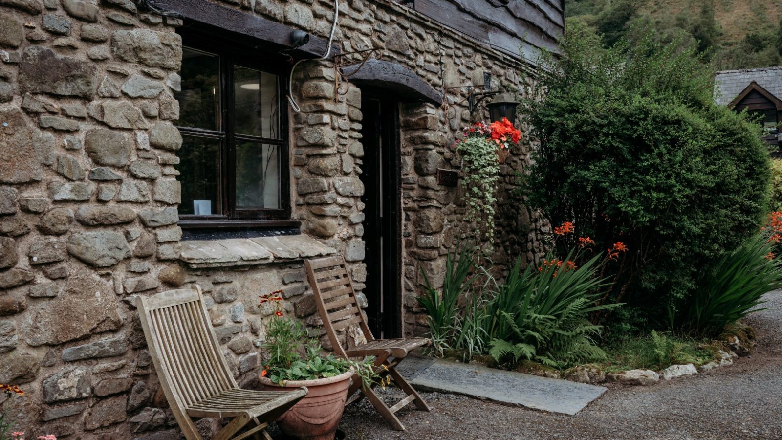 Nestled in the serene landscape of rolling hills, Cwm Chwefru Cottages features a charming stone exterior with two wooden chairs outside. Vibrant red flowers in a pot and lush greenery surround the entrance, enhancing its quaint and inviting atmosphere.