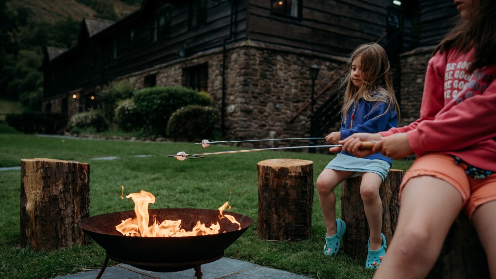 Outside the rustic stone and wood building of Cwm Chwefru Cottages, two children sit on wooden stumps, toasting marshmallows over a fire pit in the grassy area. One child wears a blue sweater and the other a red one.