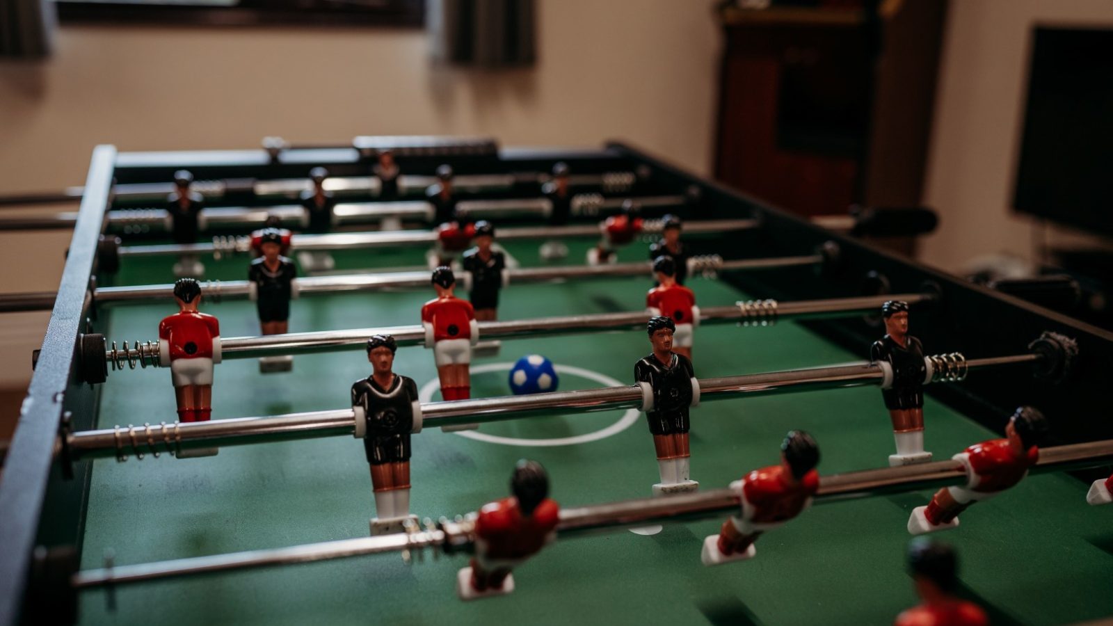 Close-up of a foosball table at Cwm Chwefru Cottages, with red and black players poised on metal rods. A small blue and white soccer ball sits on the green field. In the background, you can see blurred windows and a video game cabinet inviting guests to unwind.