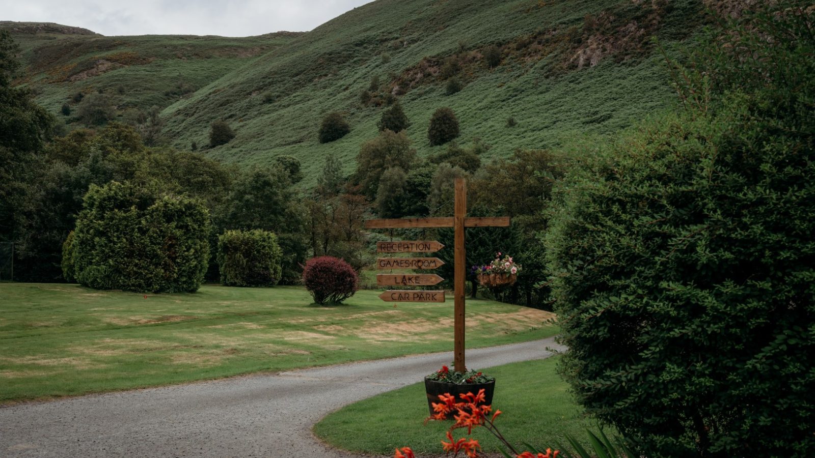 A gravel path winds through a lush, green landscape with rolling hills near Cwm Chwefru Cottages. A wooden signpost indicates directions to various locations. Bright orange flowers are in the foreground, and dense trees and shrubbery surround the area. The sky is overcast.