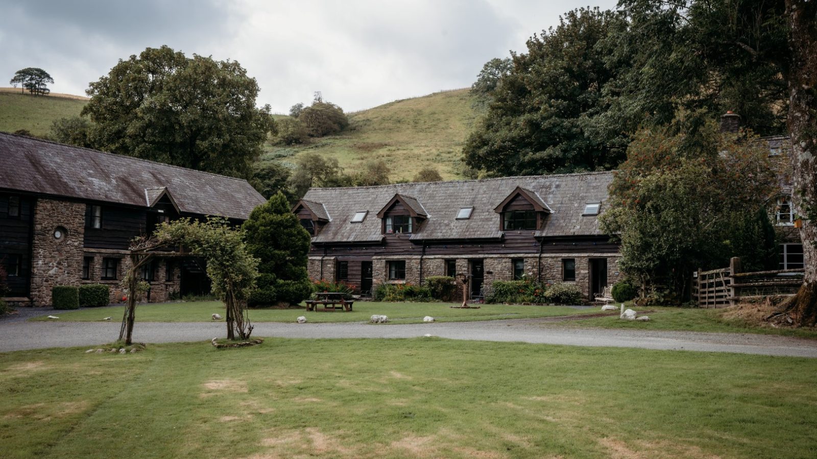 A rustic stone building with dormer windows surrounded by lush greenery, nestled in the picturesque setting of Cwm Chwefru Cottages. Situated on a grassy area with hills in the background, the cloudy sky adds a tranquil atmosphere to this charming scene.