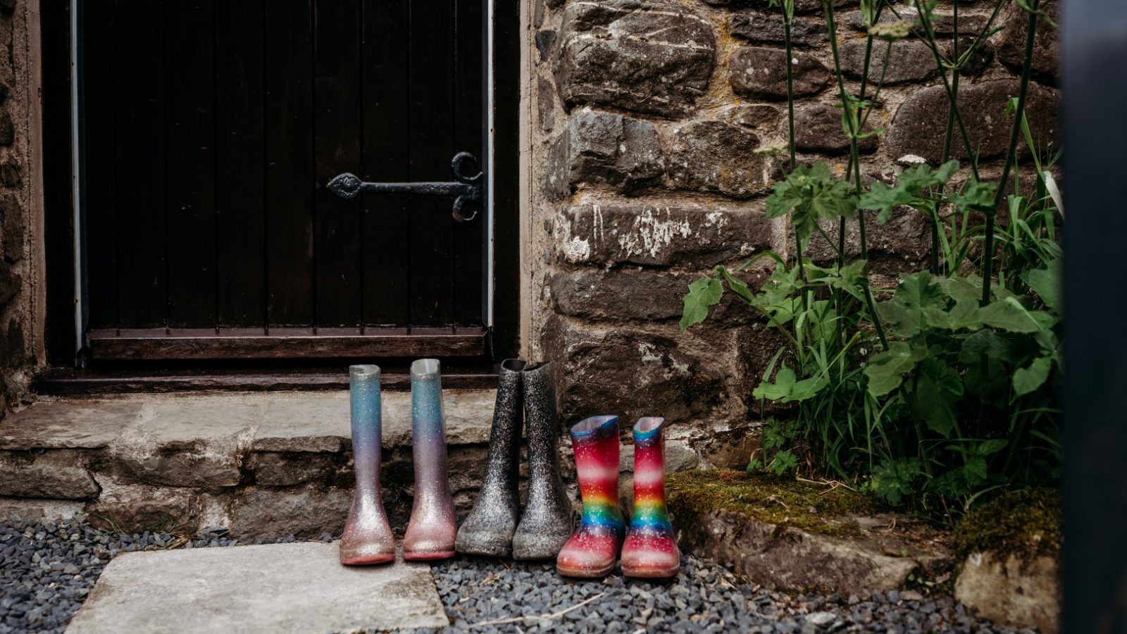 Four pairs of colorful rain boots are neatly lined up beside the wooden door of a Cwm Chwefru Cottage. Green plants grow by the stone wall, and the ground is covered with small stones.