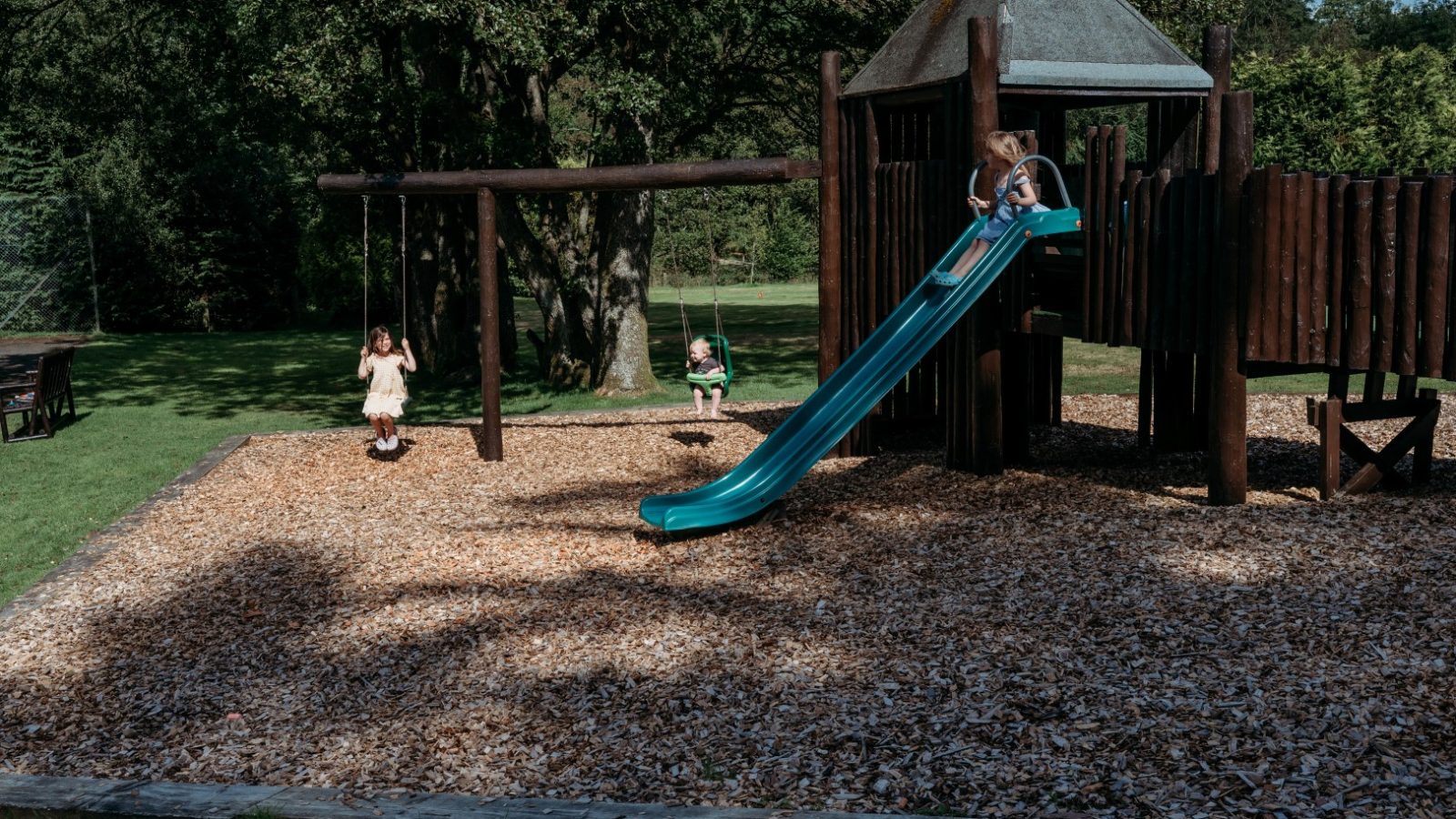 Children are playing on a playground set in a park near Cwm Chwefru Cottages. One child is on a swing, another is climbing a wooden structure, and a third is sliding down a blue slide. The ground is covered with wood chips, and trees from the surrounding cottages provide a scenic backdrop.