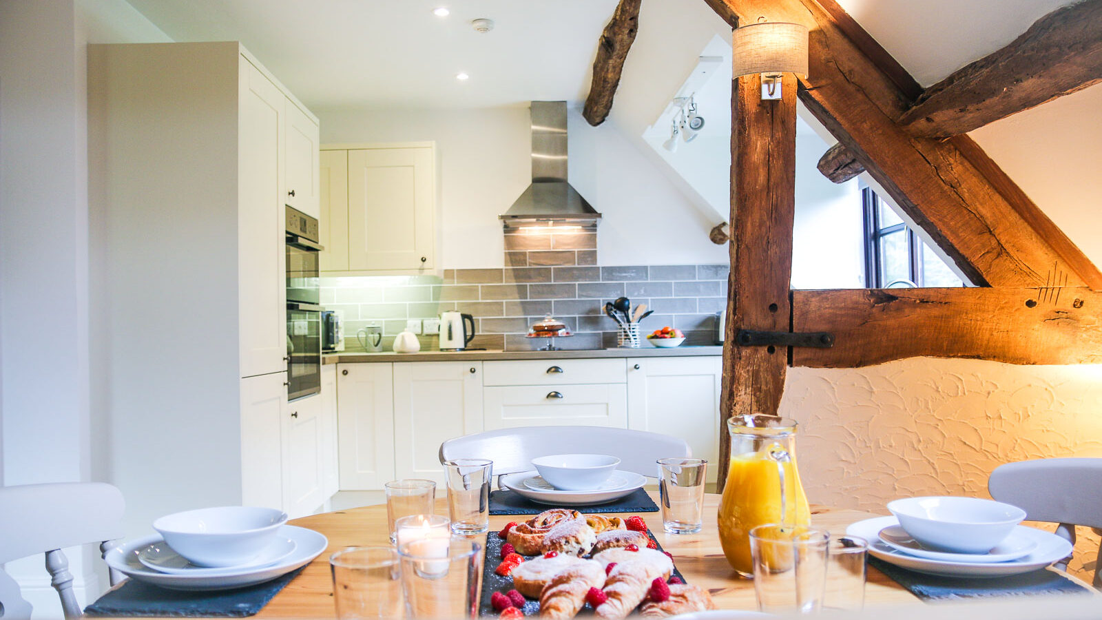 This modern kitchen in Cwm Chwefru Cottages features exposed wooden beams and a dining table set with breakfast items, including plates, glasses, and a jug of orange juice.