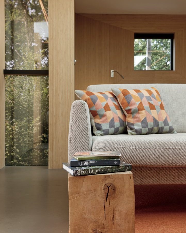 Modern living room with a gray sofa, geometric cushions, and a wooden block coffee table adorned with books. A large window frames the view, reminiscent of a Pinwheel Treehouse where nature meets style seamlessly.