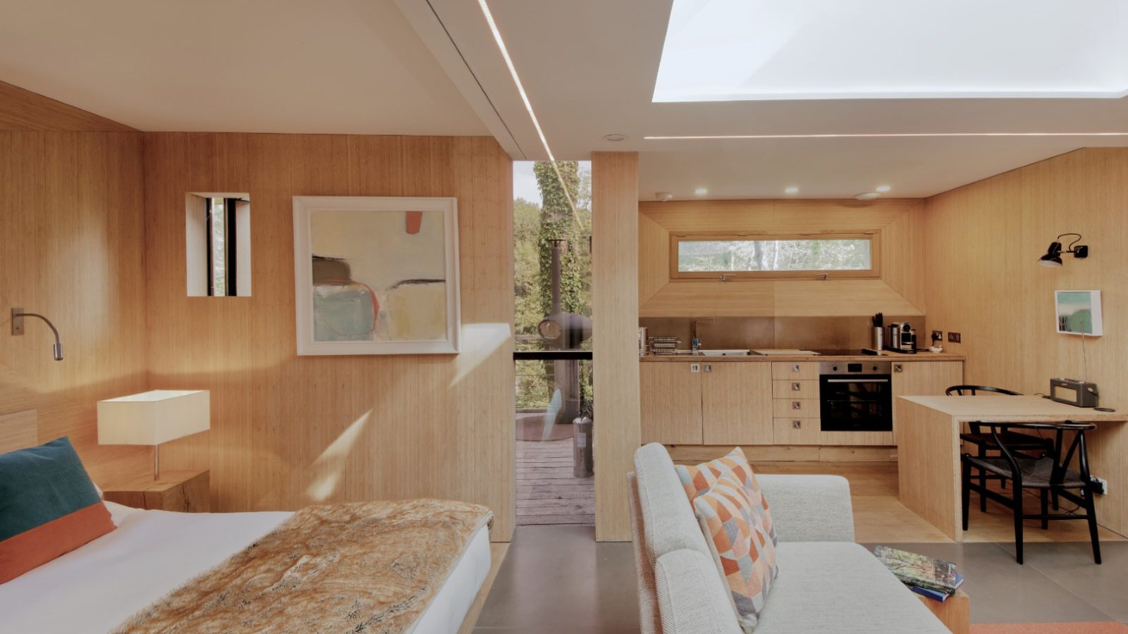 This modern, compact studio, reminiscent of a Pinwheel Treehouse, features wooden interiors, a cozy bed, sofa, kitchenette, dining area, and a skylight that bathes the space in natural light.