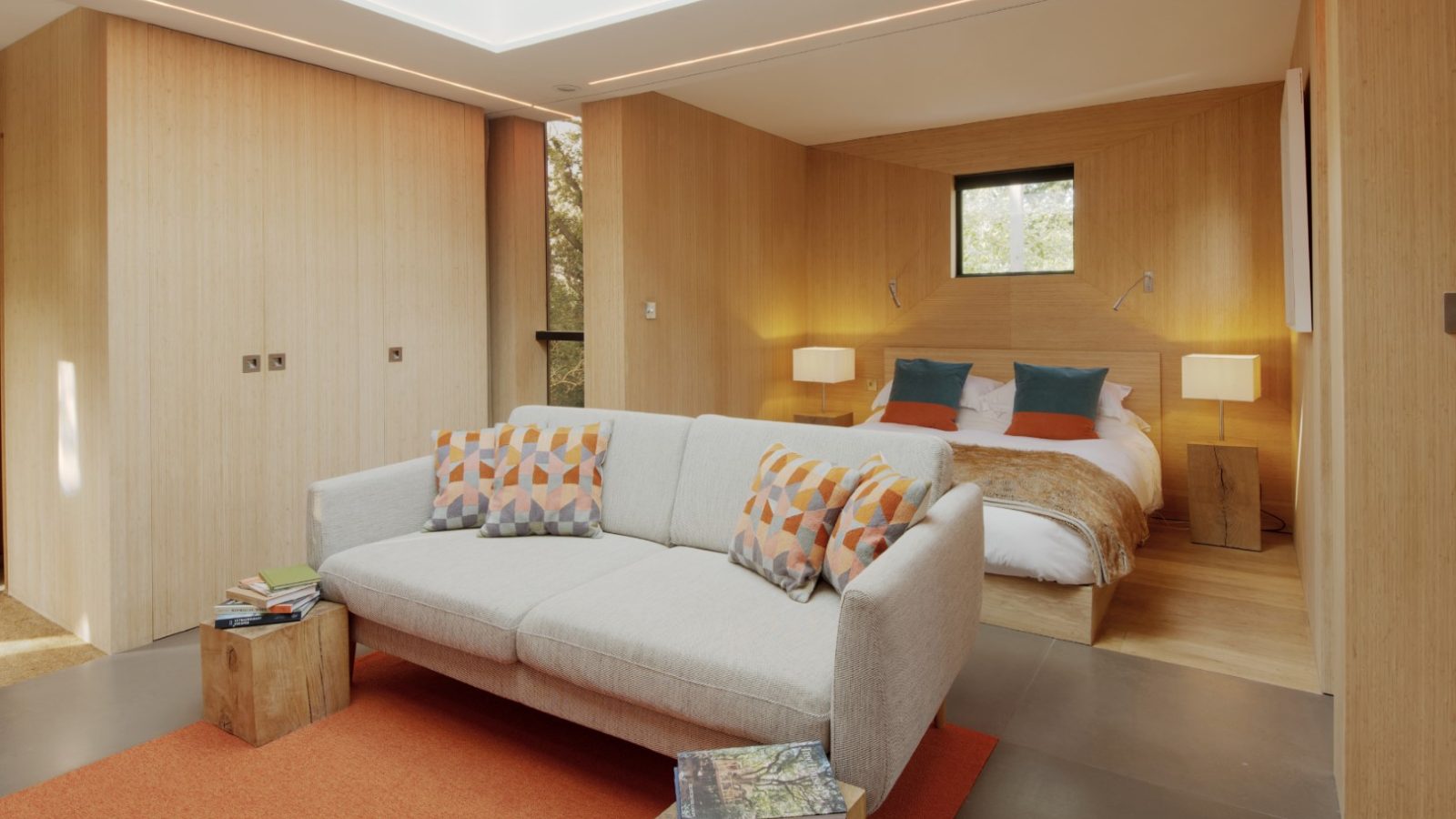 Modern hotel room with a double bed, a beige sofa adorned with colorful pillows, and wooden interior accents that create a cozy treehouse vibe.