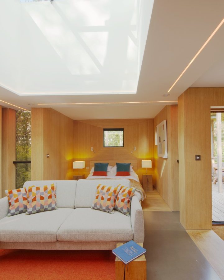 Modern treehouse bedroom with a skylight, featuring a cozy bed, sofa adorned with colorful pillows, nightstands with lamps, and an open book on a small table.