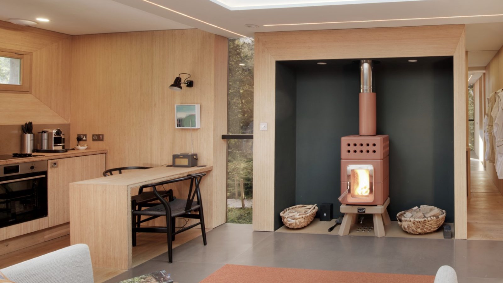 This modern Pinwheel Treehouse-themed living room features wooden walls, a wood-burning stove, a cozy kitchen area, and a seating space under a skylight for a touch of natural elegance.