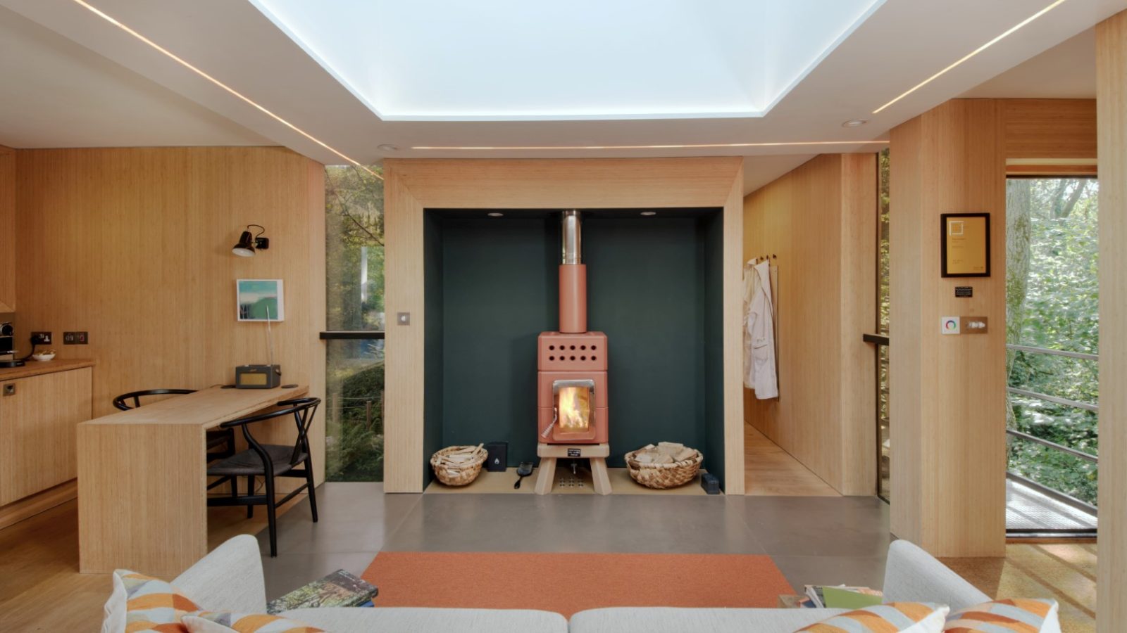 A modern treehouse-inspired living room features a cozy wood stove, skylight, and light wood furnishings. On the left side, a desk offers a serene window view that captures the essence of nature.