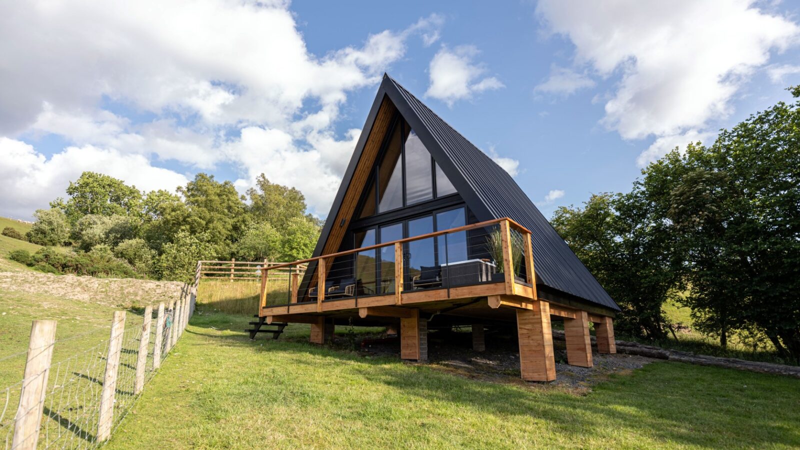 Sigrid Lodge is an A-frame cabin with large windows and a wooden deck, perfectly perched on a grassy hill surrounded by trees under a partly cloudy sky.