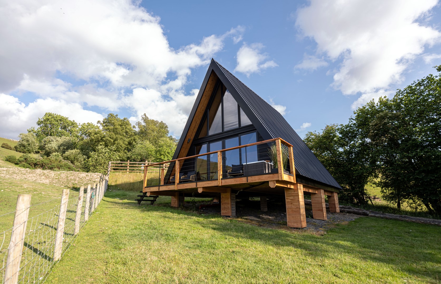 Sigrid Lodge is an A-frame cabin with large windows and a wooden deck, perfectly perched on a grassy hill surrounded by trees under a partly cloudy sky.