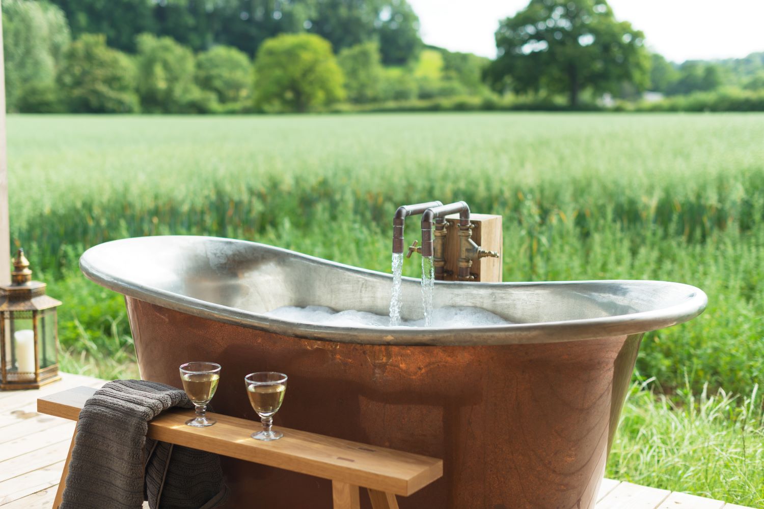 A luxurious copper bathtub is set on a deck overlooking a lush, green field. Two glasses of white wine and a folded towel rest on a wooden bench beside the tub. Water fills the tub, and the scene is serene and inviting—a cosy retreat with trees and autumnal greenery in the background.