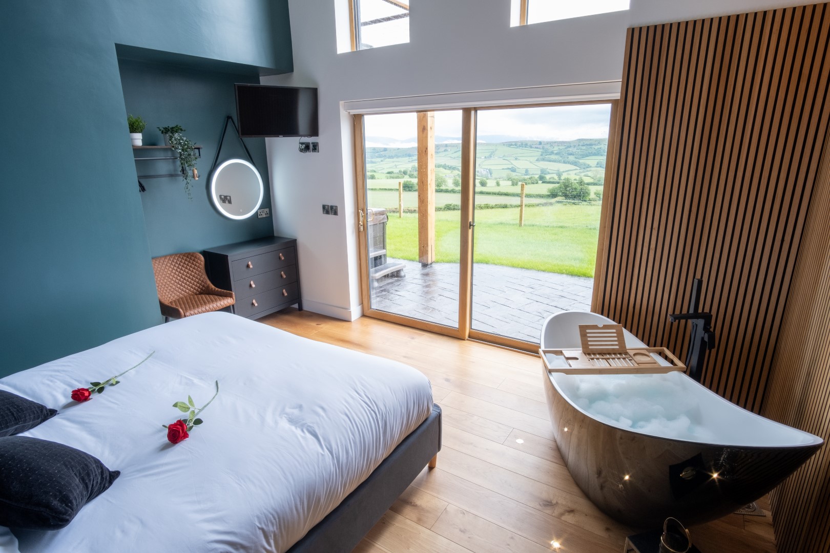 A modern, cosy bedroom with a neatly made bed adorned with two red roses. There's a comfy chair, wall-mounted TV, a round mirror over a dresser, and a luxurious bathtub filled with bubbles. Large glass doors open to an autumnal view of rolling green hills and open fields—a perfect autumn getaway from everyday life.