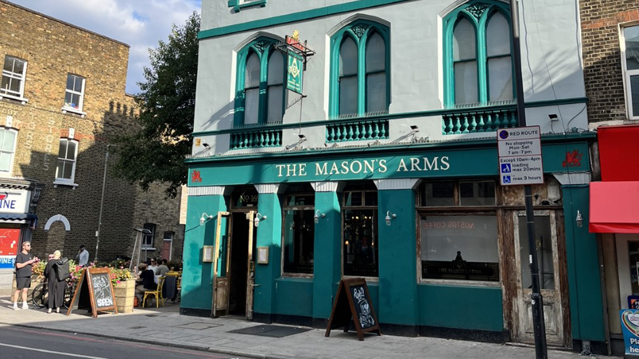 Street view of The Mason's Arms pub, a cosy autumnal retreat with its green facade, arched windows, and inviting outdoor seating. The scene unfolds under a partly cloudy sky.