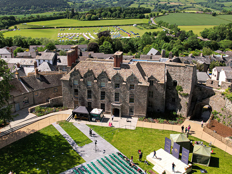 Aerial view of a historic stone building surrounded by a village and green fields, offering the perfect setting for cosy autumnal retreats, with tents visible in the distance.