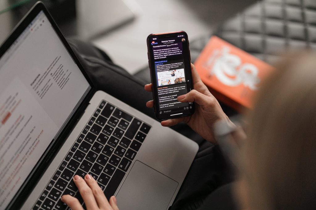 A person is using a smartphone and a laptop simultaneously. The phone displays a news article titled "Top 4 Methods of FREE Promotion For Your Holiday Home," while the laptop screen shows text and images. They are seated, with a red box in the background on a quilted surface. The scene suggests multitasking or researching.