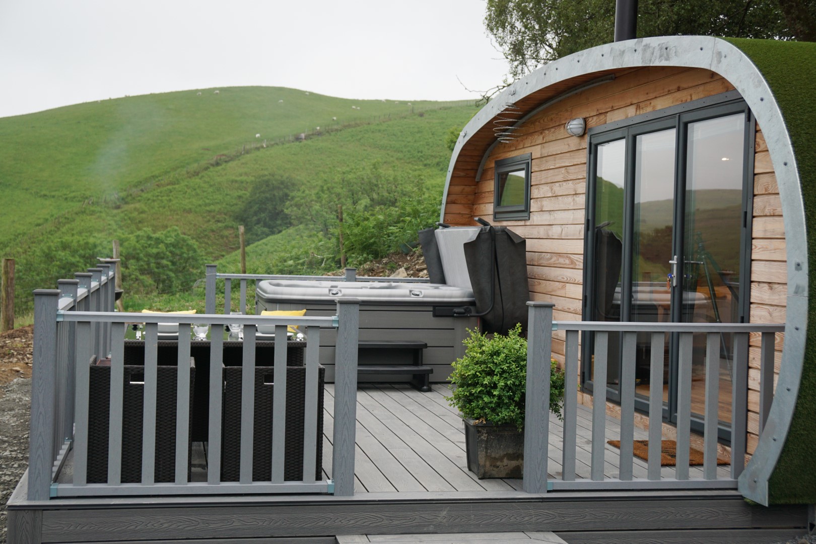 A cosy glamping cabin with a curved roof and a deck featuring a hot tub and outdoor seating, perfect for those seeking cheap breaks with hot tubs. The cabin is set on a hillside with lush green grass and rolling hills in the background. The deck is enclosed with a grey railing and includes potted plants for decoration.