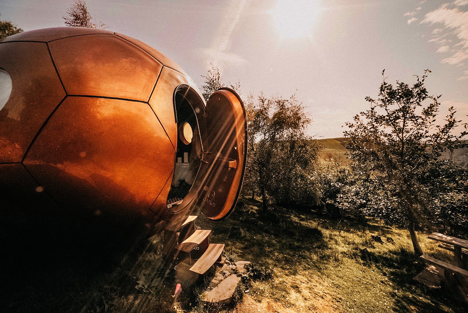 A spherical wooden cabin with an open door sits in a grassy area, surrounded by trees under a sunny sky—perfect for those seeking remote getaways in the UK.