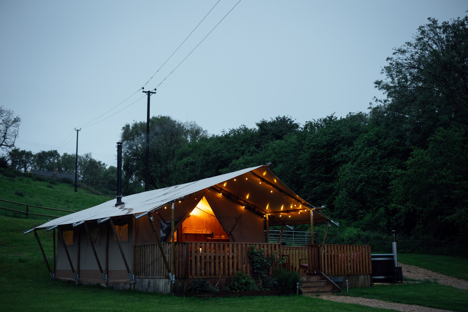 A well-lit safari tent is set up on a wooden deck in a grassy area surrounded by trees. The tent features string lights along its edges, creating a cosy and inviting atmosphere during twilight. A dirt path leads up to the tent, the perfect remote getaway in the UK